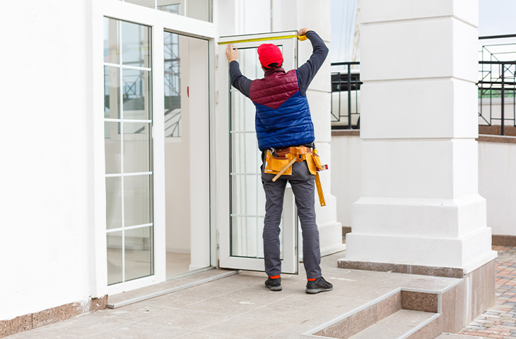 A construction worker in a red hat and tool belt measures a glass door frame with a tape measure outside a white building.
