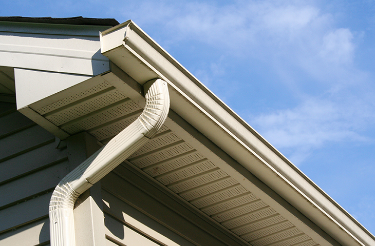 Close-up view of a house’s white eaves, soffit, and gutter system against a blue sky with light clouds. The gutter curves downward into a vertical downspout attached to the siding.