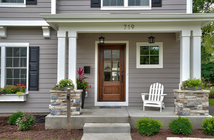 A front porch of a gray house with white trim, a wooden door, two stone columns, an Adirondack chair, hanging lanterns, flower boxes under windows, and shrubs along the walkway.