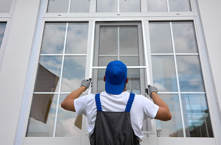 A worker in overalls and a blue cap installs or repairs a window screen on a large glass window, viewed from behind.