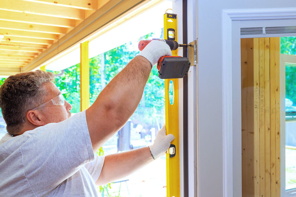 A person wearing safety glasses and gloves uses a drill and a yellow level to install a door in a wooden frame, with natural light coming through large windows in the background.