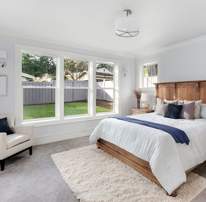 Bright bedroom with large windows, a wooden bed with white bedding and blue accents, a beige armchair, soft gray carpet, and a fluffy white rug. The room overlooks a green backyard and has simple, modern decor.