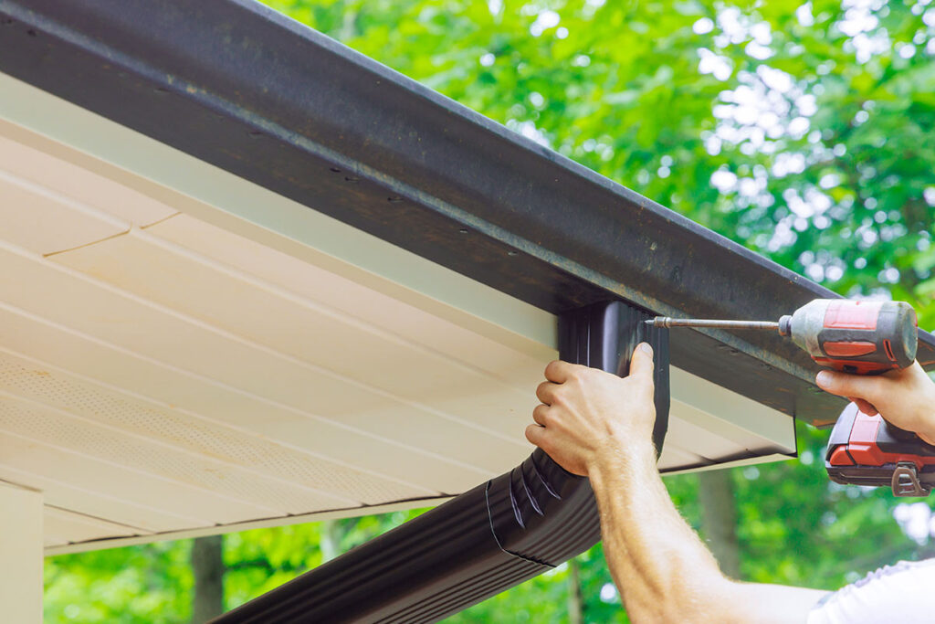 A person uses a power drill to attach a black gutter downspout to the edge of a roof, with green trees visible in the background.