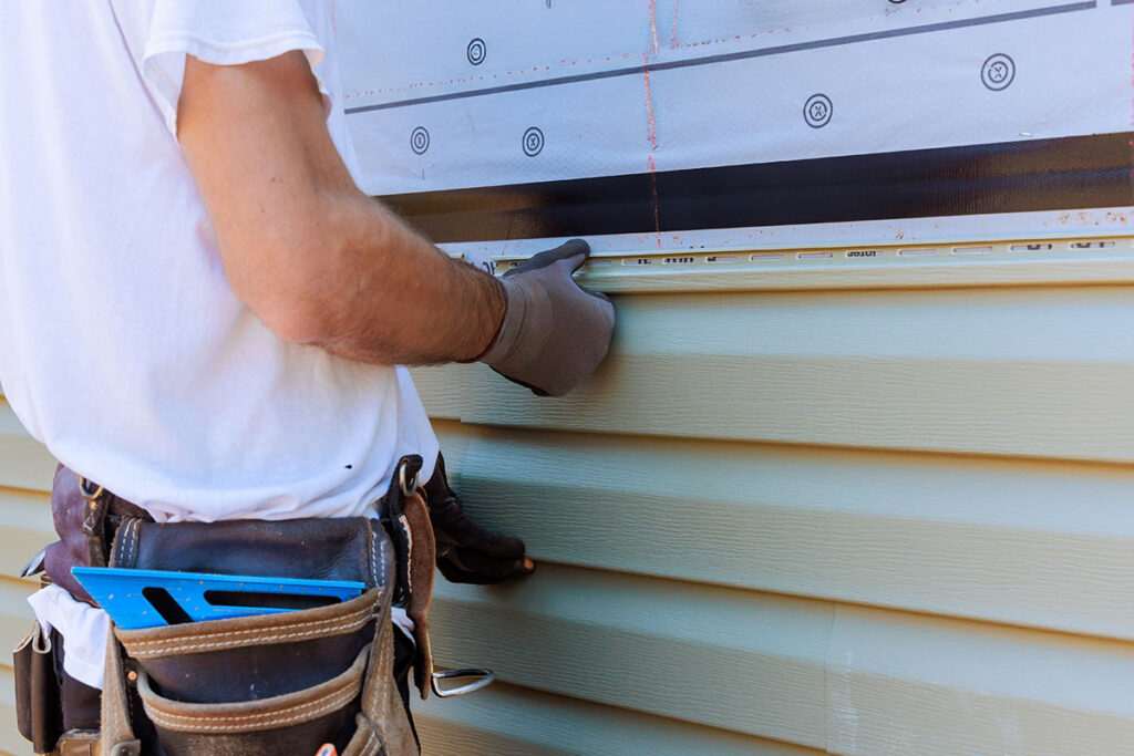 A person wearing work gloves and a tool belt installs vinyl siding on a house exterior, aligning the panel under a piece of metal trim. Only the persons torso and arms are visible.